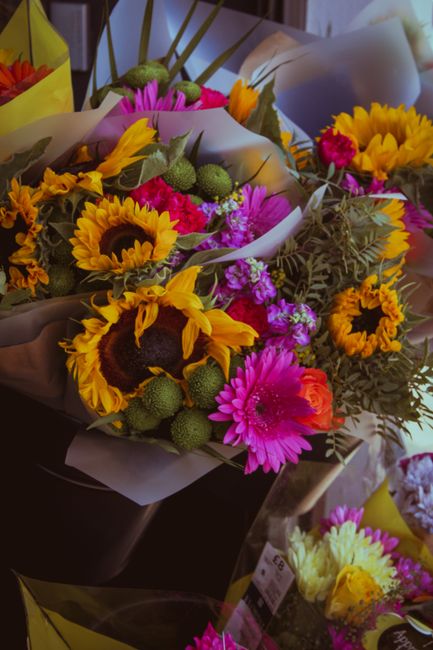 A vibrant floral bouquet arranged with large yellow sunflowers featuring dark centers, bright pink and orange dahlias, as well as smaller purple flowers, all interspersed with green foliage and round 