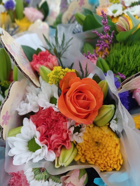 Close-up of a vibrant floral bouquet featuring an orange rose at the center, surrounded by pink carnations, white daisies, yellow chrysanthemums, and purple lavender sprigs. The flowers are wrapped in