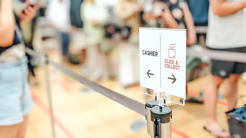 A close-up of a transparent sign with a white background displaying 'CASHIER' and directions for 'CLICK & COLLECT' with black arrow icons, situated on a black stand at the checkout counter of a busy f