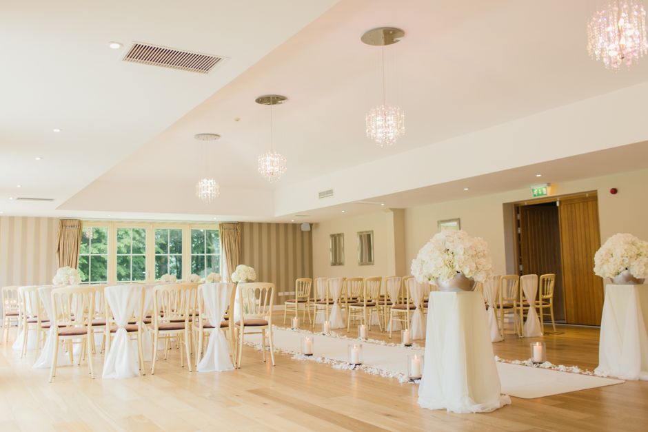 A spacious, well-lit wedding ceremony room decorated with elegant floral arrangements. Two large white vases filled with lush, white hydrangeas are positioned on tall, white pedestal tables at the fro