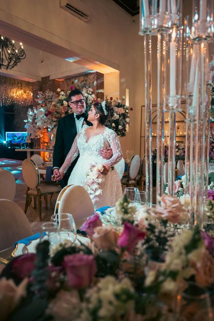 A bride and groom sharing a tender moment at their wedding reception within an elegant venue. The bride is wearing a white lace wedding gown with long sleeves, holding a bouquet of white and blush flo