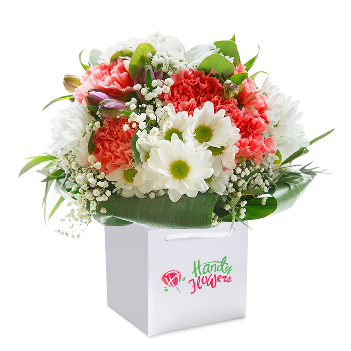 A brilliantly designed floral bouquet sits upright within a modern, square-shaped white paper gift bag, the entire setting illuminated by soft, even light and placed against a bright white backdrop. At its centre, coral-pink and vivid red carnations draw the eye with intricate, frilled petals that create a full-bodied, richly textured feel-evocative of the lush borders found in West Heath's cherished community parks. Interspersed are pure white daisies with bright chartreuse centres, providing a refreshing contrast and an uplifting mood. Delicate baby's breath branches weave between the primary flowers, their tiny white blossoms adding a filigree of gentle grace. Tucked in at the edge, a handful of unopened buds washed in purple and rose add intrigue and gentle movement, reminiscent of morning walks along West Heath Road, where new blooms unfurl daily. The base and perimeter are surrounded by wide, glossy green leaves that gather the arrangement in an organic embrace. The white bag, topped with sturdy ribbon handles, stands ready for easy gifting or local delivery. This expertly arranged bouquet perfectly expresses the spirit of West Heath's floral artistry, making it an excellent choice for birthdays, memorable anniversaries, or simply to convey sincere appreciation delivered to a loved one nearby.