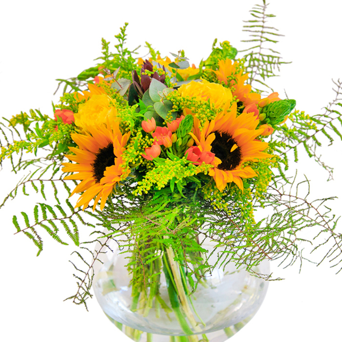 An effusive burst of summer warmth, this bouquet greets the eye with the radiant sheen of sunflowers and the soft glow of golden garden roses, carefully curated by a West Heath florist steeped in local tradition. Each sunflower's bold, velvet center stands proud, surrounded by wide petals the color of midday sunshine; beside them, yellow roses unfurl in precise, multilayered spirals, offering a plush, elegant counterpoint. Scarlet hypericum berries dot the design, their rich, luminous hue a lively accent amid the greenery's gentle tones. Cascading over the vase's edge, feathery fern fronds and mists of airy goldenrod echo the wild beauty of West Heath's ancient woodland boundaries, while slender spikes of light green foliage and bluish eucalyptus add both structure and rustic character. The round, transparent glass vase cradles every stem, water gleaming in natural daylight, emphasizing the bouquet's vibrant, clean lines. Up close, the fragrant scent of roses and eucalyptus mingles softly, suggesting the freshness of West Heath gardens at their peak. Ideal for birthdays, anniversaries, or simply celebrating life's joyful milestones, this bouquet captures the spirit and beauty of the local community-bringing the essence of a West Heath summer into any home.
